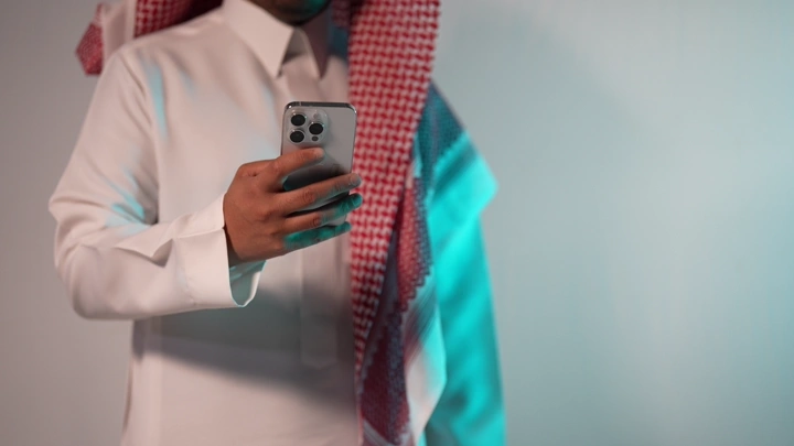 رجل سعودي يرتدي ثوبًا وغترة يمسك هاتفًا ذكيًا، لقطة داخلية.