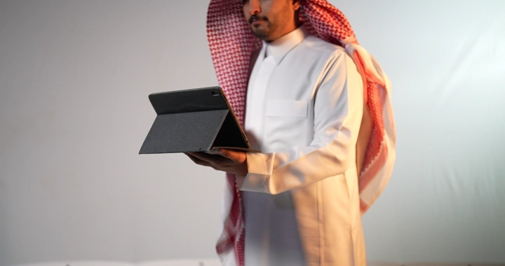 رجل سعودي يرتدي ثوبًا وغترة يحمل جهاز آيباد، لقطة داخلية.