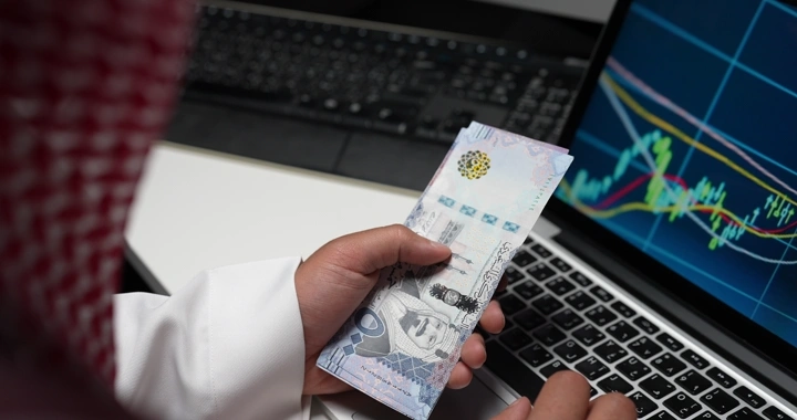 رجل سعودي يعد النقود أمام لابتوب يعرض مخططًا بيانيًا، لقطة مقربة.