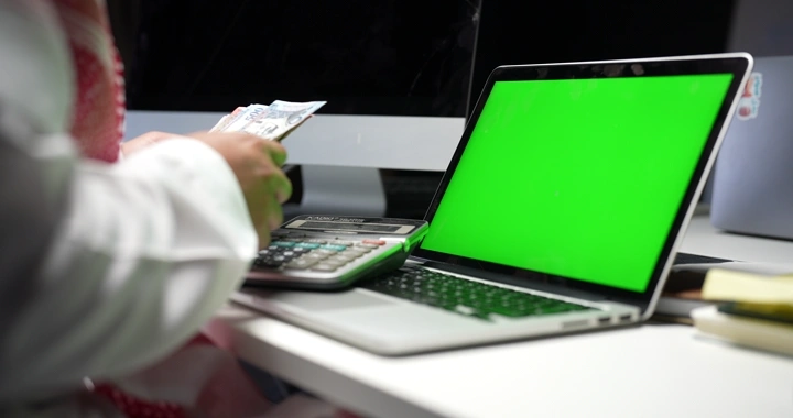 رجل سعودي يعد المال بجانب حاسوب محمول بشاشة خضراء، لقطة داخلية.