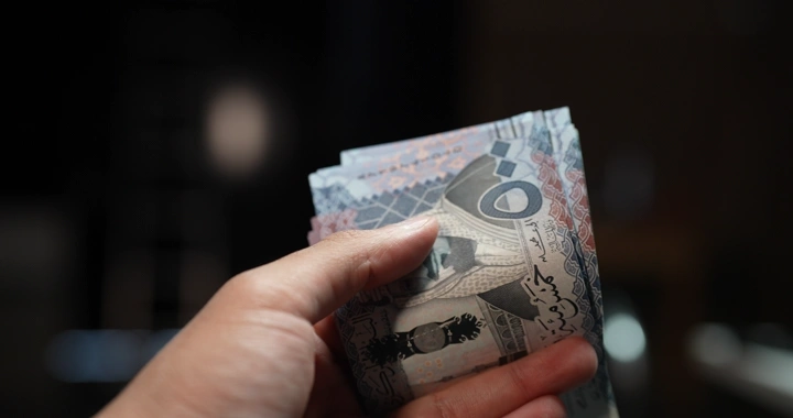 يد تمسك بأوراق نقدية سعودية في إضاءة خافتة، لقطة قريبة.
