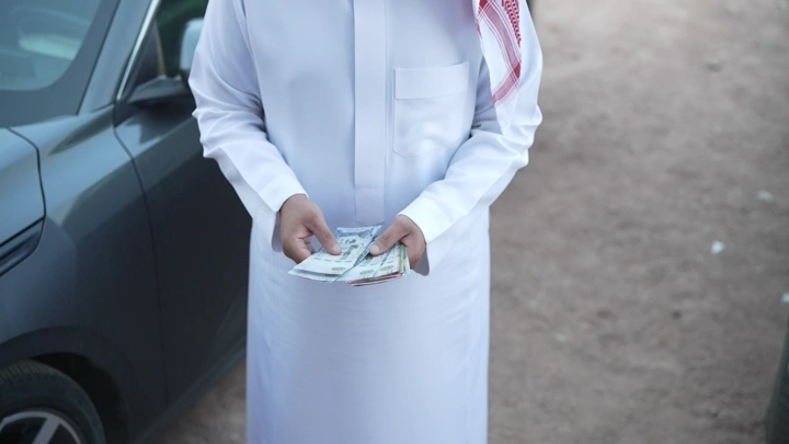 رجل يرتدي ثوبًا يعد النقود بجانب سيارة، لقطة قريبة.