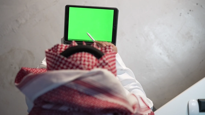 رجل سعودي يستخدم جهاز لوحي بشاشة كروما خضراء، لقطة من الخلف.