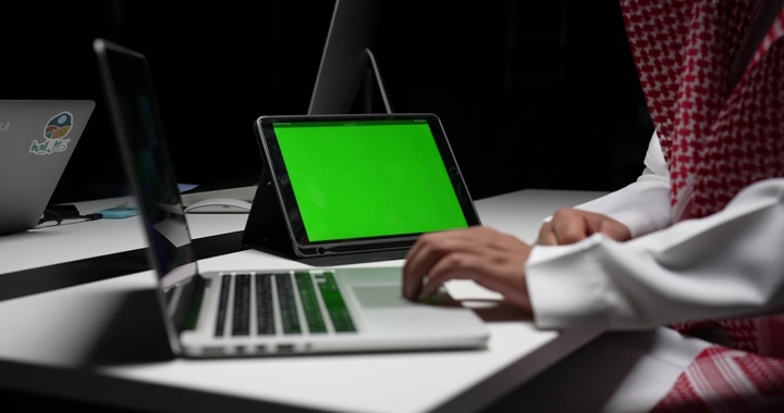 رجل سعودي يعمل على جهاز كمبيوتر محمول أمام شاشة كروما خضراء، لقطة داخلية.