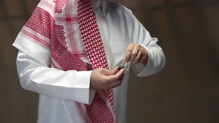 رجل يرتدي ثوباً وغترة يمسك بزجاجة دهن عود، لقطة قريبة.