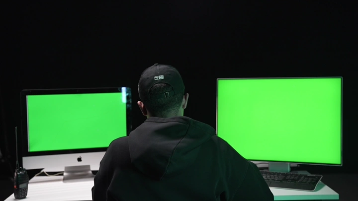 رجل يجلس أمام شاشات خضراء في غرفة مظلمة، لقطة من الخلف.