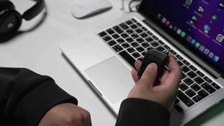 يد تمسك جهاز تحكم صغير أمام لابتوب على مكتب، لقطة مقربة.