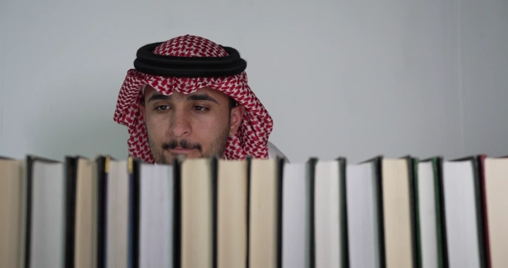 رجل يرتدي شماغ يقرأ كتابًا أمام رف كتب، لقطة أمامية.