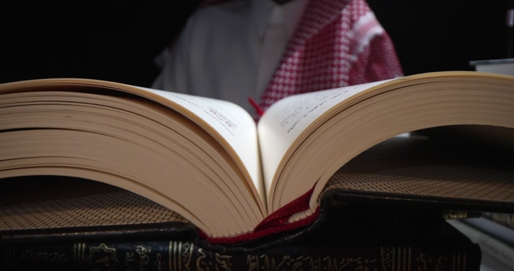 كتاب مفتوح على طاولة مع رجل يرتدي شماغ في الخلفية، لقطة قريبة.