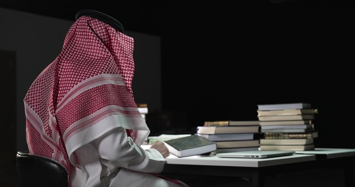 رجل يرتدي الزي التقليدي يقرأ بين الكتب في غرفة مظلمة، لقطة جانبية.