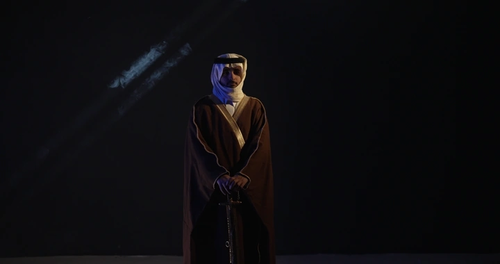 رجل يرتدي الزي التقليدي واقفًا حاملاً سيفًا، إضاءة خافتة.