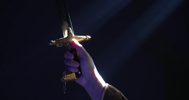 يد ترفع سيفًا ذهبيًا في ضوء خافت، لقطة مقربة.