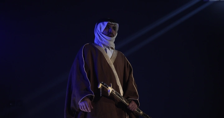 رجل يرتدي ثوباً تقليدياً يحمل سيفاً في إضاءة سينمائيه.