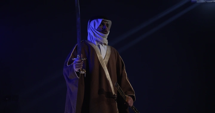 رجل يرتدي زي تقليدي يرفع سيفه في إضاءة سينمائيه.