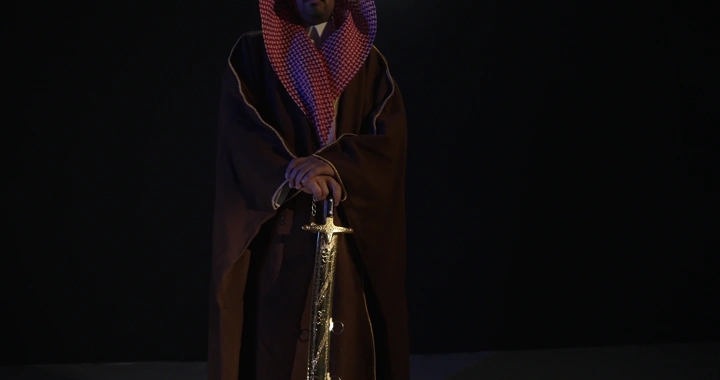 رجل يرتدي ثوبًا تقليديًا ممسكًا بسيف، إضاءة منخفضة.