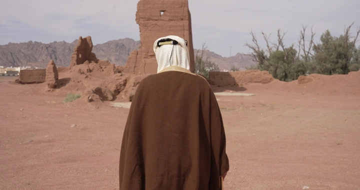 رجل يرتدي ثوبًا تقليديًا يمشي بين بيوت طينية في الصحراء، لقطة خلفية.