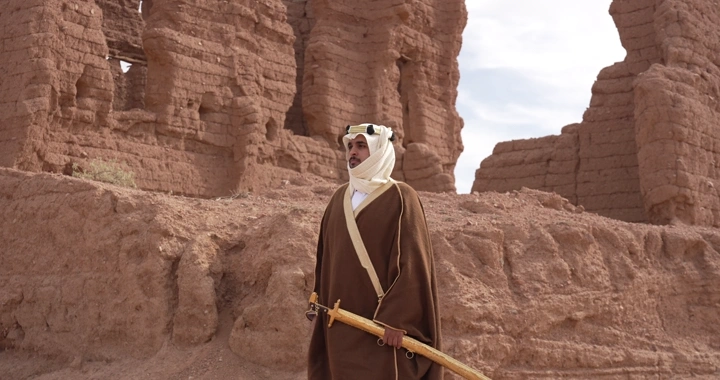 رجل يرتدي الزي التقليدي يقف أمام بيوت طينية قديمة، لقطة نهارية.