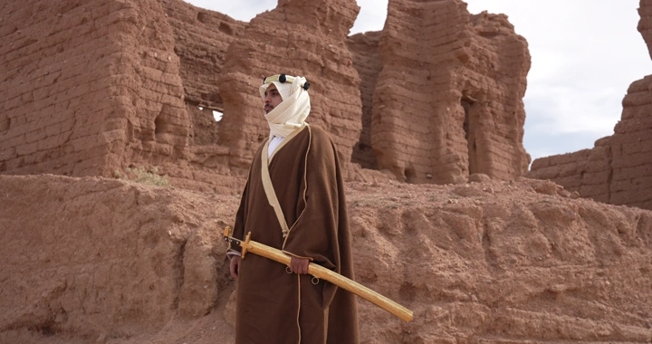 Man in traditional attire standing among mud houses, daytime shot.