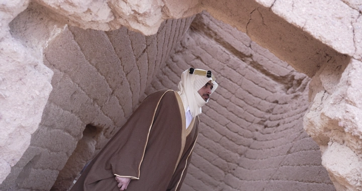 رجل يرتدي الزي التقليدي يقف بين بيوت طينية، لقطة قريبة.