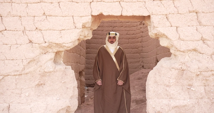 رجل يرتدي ثوبًا تقليديًا يقف بين بيوت طينية، لقطة أمامية.