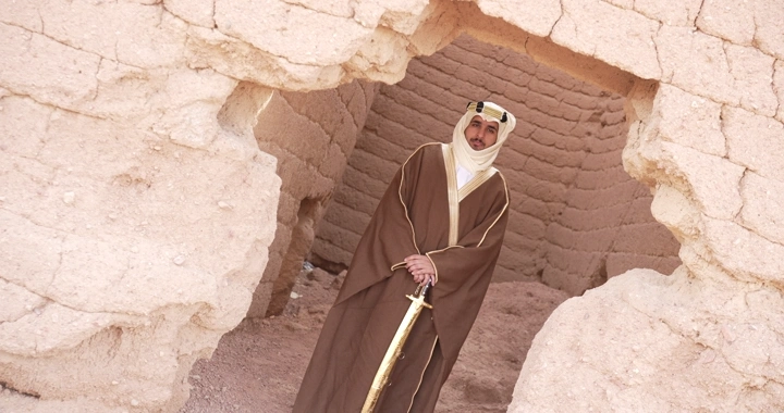 رجل يرتدي زي تقليدي يقف بين بيوت طينية، لقطة من الأمام.