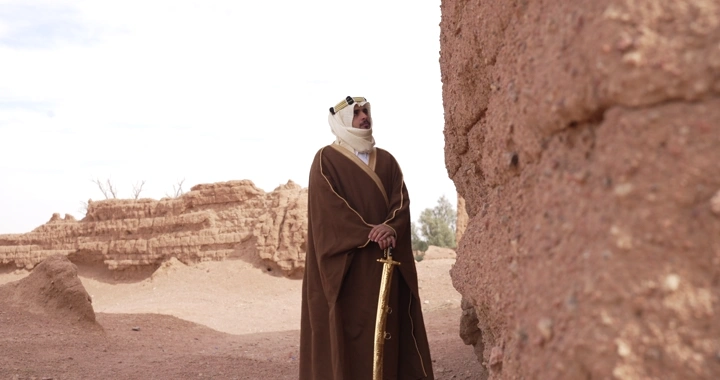 Man in traditional robe stands between mud houses, side shot.