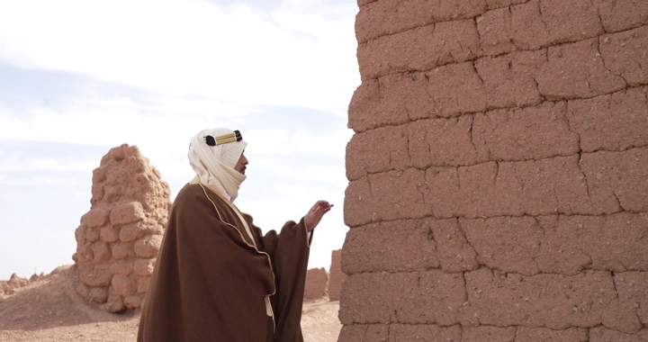 رجل يرتدي عباءة واقف بجانب بيت طيني في الصحراء، لقطة جانبية.