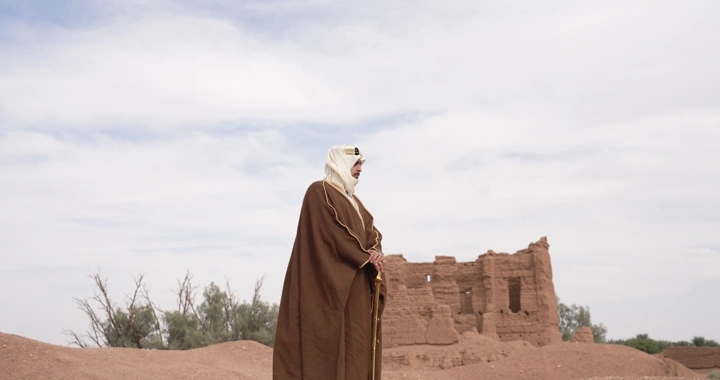 A man wearing a cloak standing among mud houses, daytime shot.
