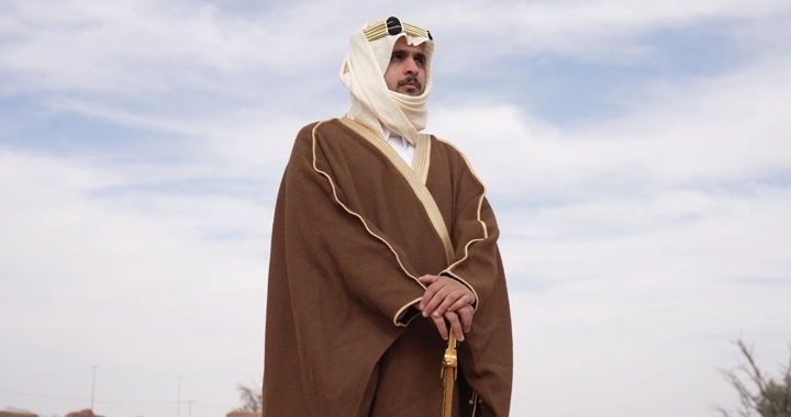 Man in traditional attire standing in a desert area, eye-level shot.