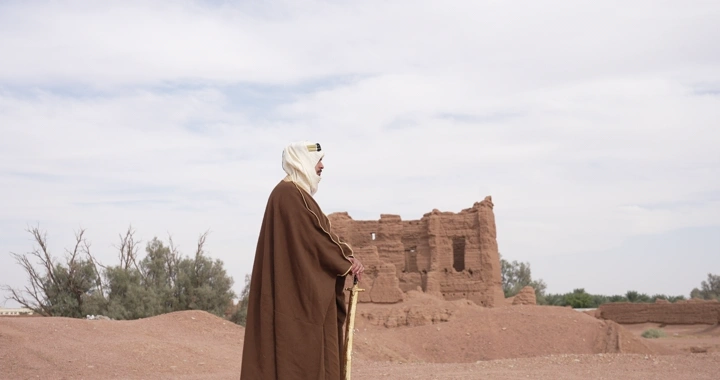 Man in robe standing before mud houses, side shot.