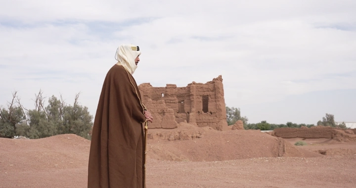 رجل يرتدي عباءة واقف بين بيوت طينية، لقطة جانبية.