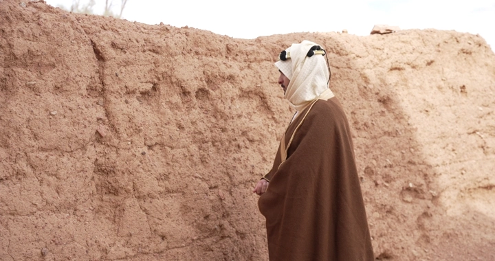Man in traditional robe standing by mud wall, side shot.