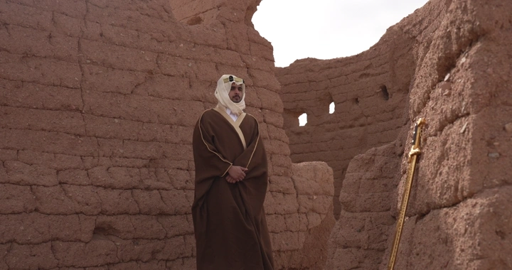 رجل يرتدي الزي التقليدي يقف بين بيوت طينية، لقطة نهارية.