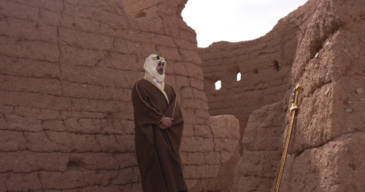 رجل يرتدي الزي التقليدي يقف بين بيوت طينية، لقطة ثابتة.