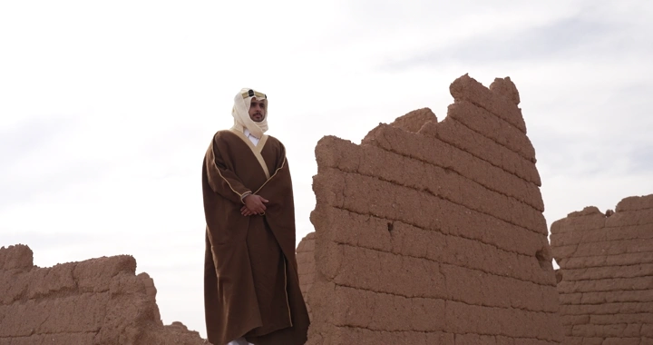 رجل يرتدي ثوباً تقليدياً يقف بين بيوت طينية، لقطة نهارية.