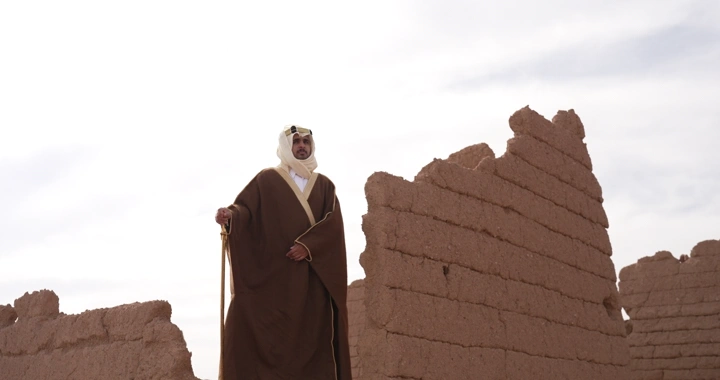 رجل يرتدي عباءة واقف بين بيوت طينية، لقطة من الأسفل.