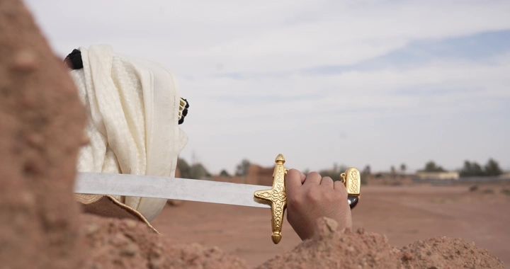 رجل يحمل سيفًا بين بيوت طينية في الصحراء، لقطة جانبية.