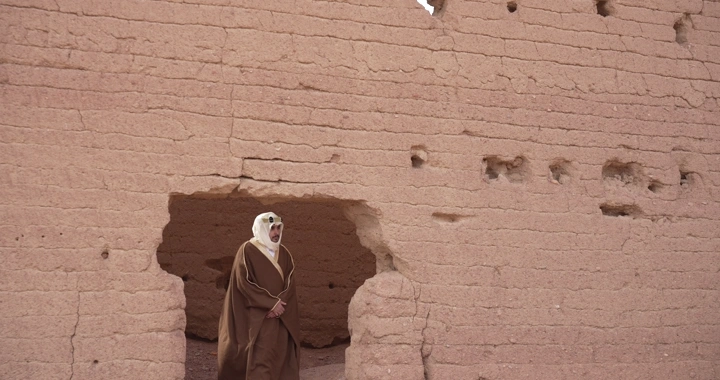 رجل يرتدي ثوبًا تقليديًا يقف بين بيوت طينية، لقطة قريبة.