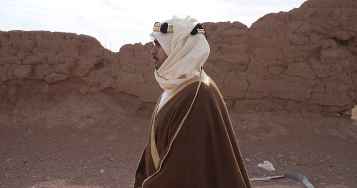 Man in traditional attire walking beside mud wall, side shot.