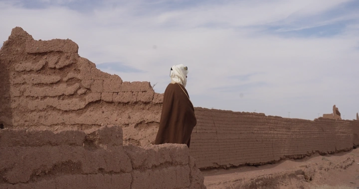 رجل يرتدي عباءة بين بيوت طينية تحت سماء غائمة، لقطة جانبية.