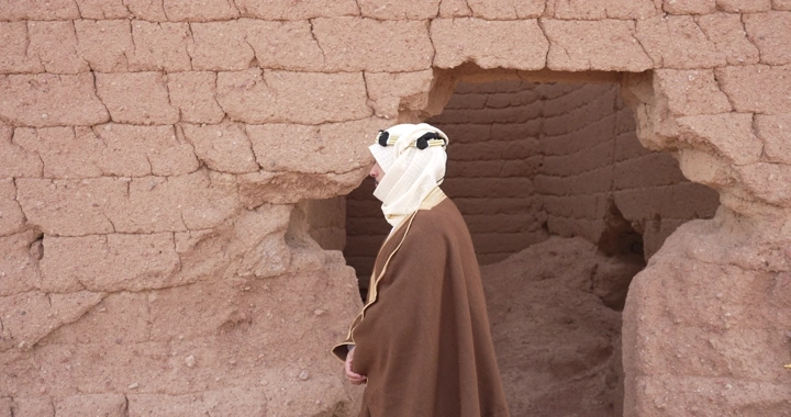 Man in traditional attire walking by mud brick wall, side shot.