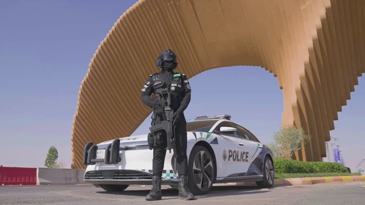 شرطي بجانب سيارة شرطة أمام هيكل معماري، لقطة نهارية.