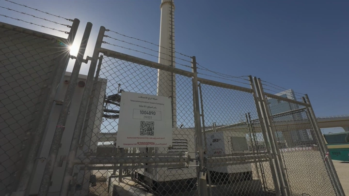 محطة كهرباء خلف سياج معدني تحت أشعة الشمس مع مهندس يصور الموقع، لقطة نهارية.