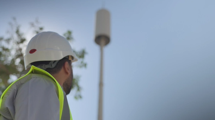 Engineer wearing helmet and safety vest looking at communication tower, side shot.