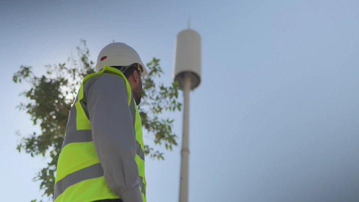 مهندس يرتدي سترة صفراء وخوذة ينظر إلى برج اتصالات، لقطة من الأسفل.