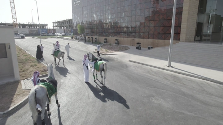 رجال بالزي التقليدي مع خيول أمام مبنى حديث، لقطة جوية.