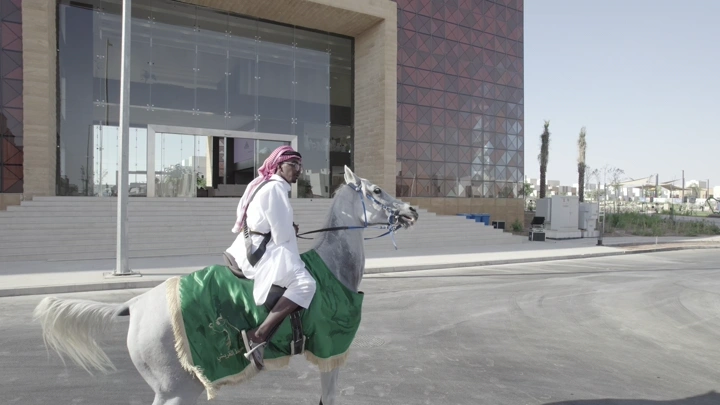 رجل يرتدي زي تقليدي يركب حصانًا أمام مبنى حديث، لقطة نهارية.
