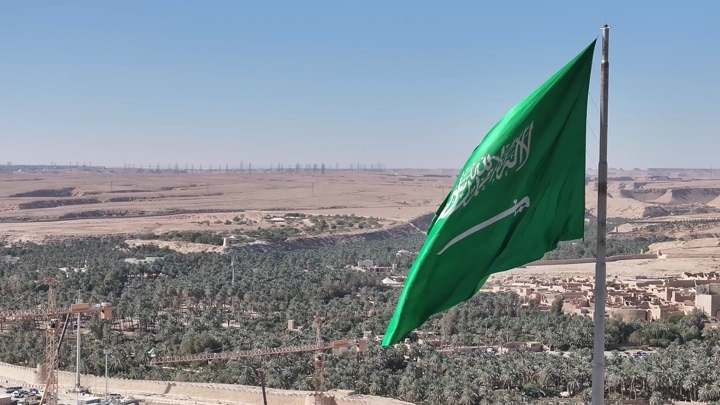 علم السعودية يرفرف فوق الدرعية في يوم مشمس، لقطة من زاوية مرتفعة.