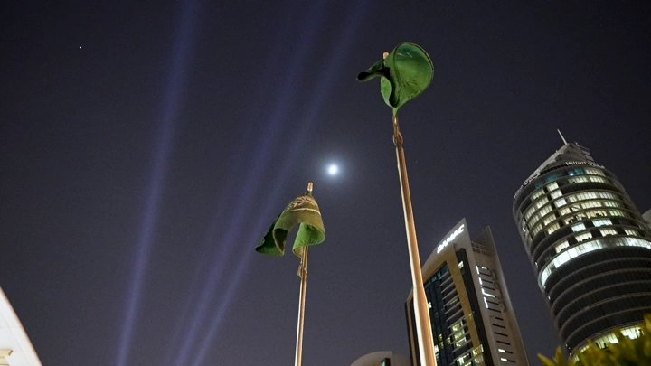 أعلام سعودية ترفرف في الليل مع أضواء المدينة، لقطة من الأسفل.