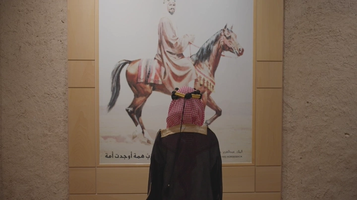 رجل يرتدي الزي التقليدي ليوم التاسيس أمام لوحة في قصر المصمك، لقطة داخلية.